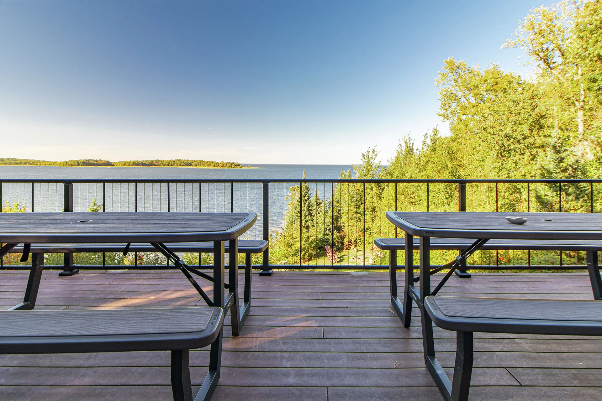 Two picnic tables on a wooden deck overlooking a lake with evergreen trees and mountains in the distance