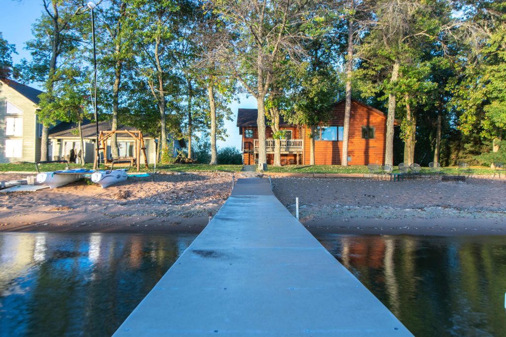 Blue boardwalk extending over water toward playground equipment and buildings surrounded by tall trees